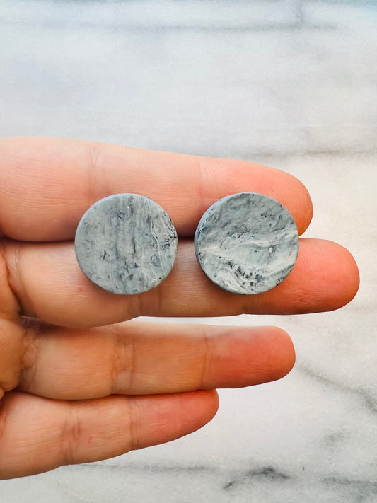 Two round, gray stone earrings held between fingers on a marble surface.